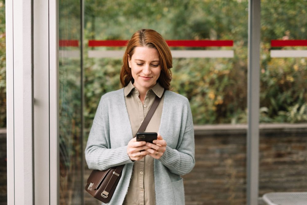 Eine Frau steht an einer Bushaltestelle und benutzt ihr Smartphone Kununu